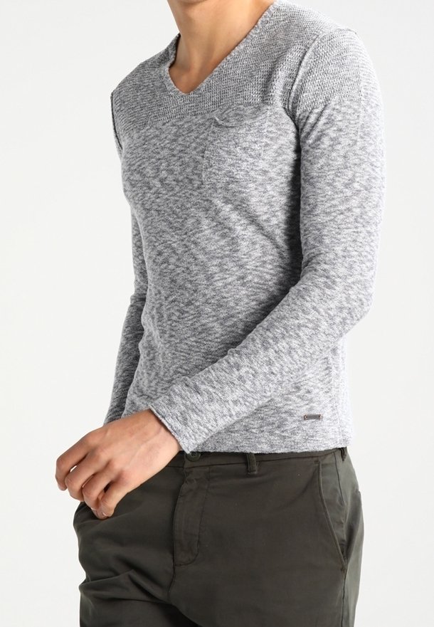 Personne portant un haut en maille gris clair ajusté à manches longues avec une petite poche poitrine et un pantalon vert foncé, debout devant un fond uni.
