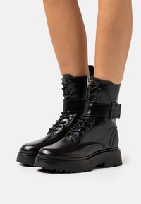 Botas negras de cuero hasta el tobillo con suela gruesa, cordones, cremallera lateral y una correa con detalle de hebilla. Textura suave con un diseño elegante.