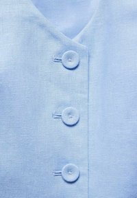 Svetle modrý lněný oděv s výstřihem do "V" a čtyřmi kulatými knoflíky potaženými látkou podél přední části. Hladká textura a čisté švy.