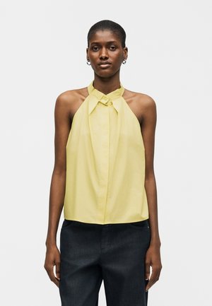 Femeie care poartă un top halter galben pal fără mâneci, cu un guler înalt și pantaloni închiși la culoare, stând în fața unui fundal simplu și deschis la culoare.