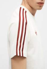 Gros plan d'une personne portant une chemise blanche à manches courtes avec trois bandes rouges sur l'épaule et un petit logo rouge sur la poitrine.