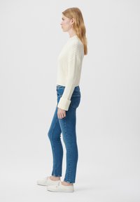 Weißer gerippter Pullover, enge blaue Jeans und weiße Sneakers. Seitenansicht vor einem schlichten Hintergrund, zeigt Slim Fit und aufgeschlagene Bündchen.