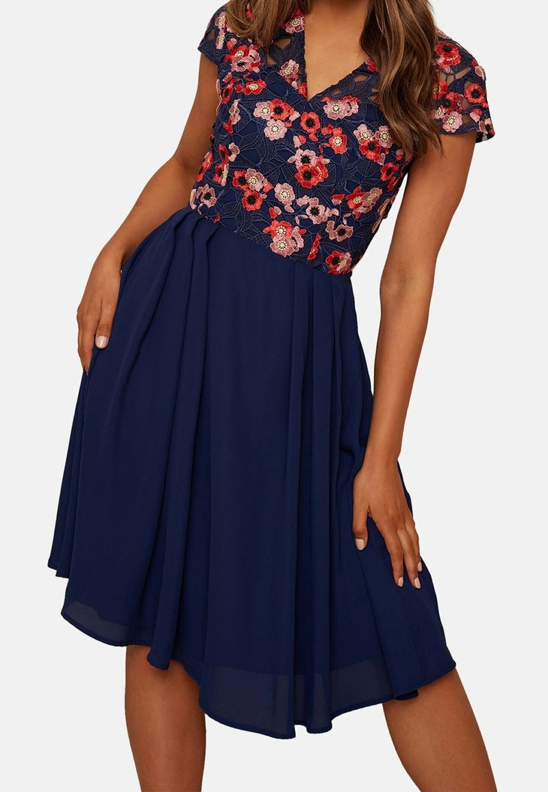 Femme portant une robe bleu marine à manches courtes et un corsage en dentelle brodée florale dans des tons rouges et roses.