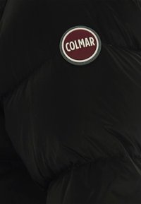 Giacca nera imbottita con una struttura liscia e una toppa rotonda, bianca e bordeaux, etichettata "COLMAR" nella zona del petto.