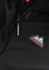 Fekete dzseki bordázott textúrával, cipzáros elöl, tépőzáras részletekkel és többszínű "SUPERDRY" logóval.