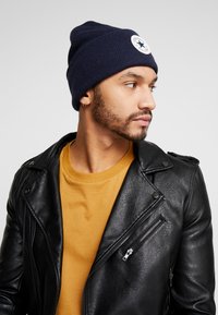 Bonnet côtelé bleu marine avec un patch rond blanc, porté avec une veste en cuir noir et une chemise moutarde. Vue de profil en gros plan.