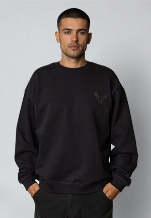 Young man with short dark hair wearing a black sweatshirt with a subtle moose antler logo on the chest, hands in pockets, neutral background.