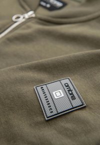 Maglietta verde oliva con scollatura con zip e un'etichetta logo nera texturizzata sul petto, con lettering in rilievo e accenti geometrici.
