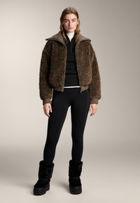 Bruine, pluizige jas met een hoge kraag, zwarte gebreide top, zwarte leggings en zwarte laarzen met bontvoering. Eenvoudige, gestructureerde stofdetails overal.