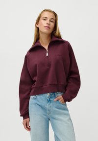 Woman with long blonde hair wearing a plum zip-up sweatshirt and light blue high-waisted jeans, hand in pocket, against a plain white background.