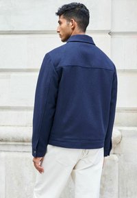 Navy blue wool blend jacket with a collar, long sleeves, and buttoned cuffs, paired with light-colored pants. Textured finish with back seam detail.