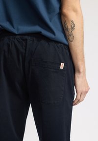 Pantaloni elasticizzati blu con un taschino posteriore dotato di etichetta. Il materiale sembra morbido con una texture liscia. Tatuaggio visibile sul braccio.