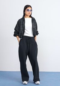 Una persona se mantiene de pie con confianza, vistiendo una chaqueta bomber negra con estampado floral, una camiseta blanca y pantalones de chándal negros, con gafas de sol azules y zapatillas blancas.