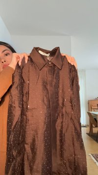 Donna che tiene una camicia marrone lucida a maniche lunghe con colletto e piccoli puntini bianchi in una stanza luminosa.