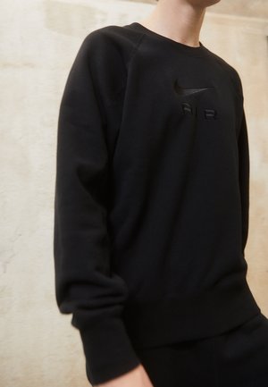 Personne portant un sweat-shirt noir avec un discret swoosh Nike noir et le mot « AIR » sur la poitrine, sur un fond clair uni.