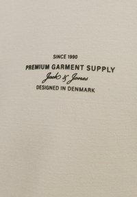 Krém színű, sima textúrájú szövet, fekete nyomtatott szöveggel, amely tartalmazza a márkanevet, valamint a tervezés és az eredet részleteit.