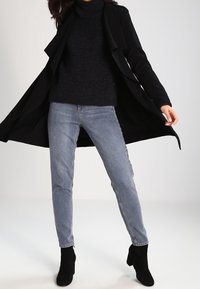 Abrigo largo negro, jersey negro grueso de cuello alto, jeans de mezclilla azul claro y botas negras hasta el tobillo. El abrigo tiene un diseño fluido y solapas.