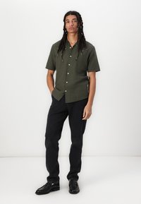 Short-sleeve, olive green button-up shirt with a relaxed fit, paired with black jeans and black leather shoes, standing on a white backdrop.