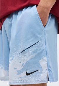Άτομο που φοράει ανοιχτό μπλε σορτς Nike με λευκό σχέδιο πιτσιλιάς και μαύρο λογότυπο, το χέρι ακουμπά στην πλαϊνή τσέπη, σε συνδυασμό με κόκκινο πουκάμισο.