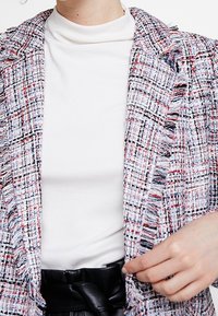 Chaqueta texturizada multicolor con un patrón de tweed en blanco, rojo y negro. Cuenta con detalles de flecos y un cuello con muesca.