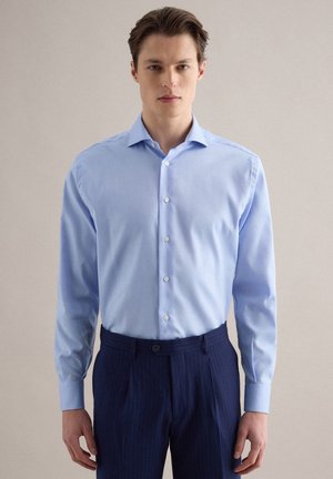 Junger Mann trägt ein hellblaues Hemd mit Knöpfen, das in dunkelblaue Nadelstreifenhose gesteckt ist, steht vor einem einfarbigen Hintergrund.