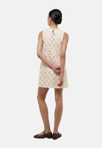 Robe sans manches crème avec un motif en dentelle florale, détail en forme de trou de serrure dans le dos, et une coupe droite, associée à des chaussures marron.