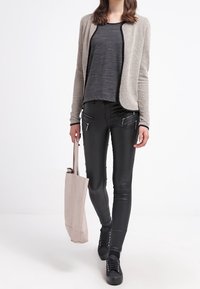 Cárdigan texturizado beige sobre una blusa negra, combinado con pantalones ajustados de piel sintética negra. Zapatillas negras y un bolso tote de color beige claro.