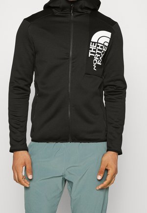 Homme portant un sweat à capuche zippé noir avec le logo "The North Face" sur la poche poitrine et un pantalon bleu clair, debout devant un fond uni.