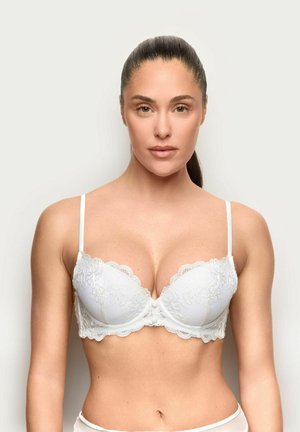 Kobieta z ciemnymi włosami w białym koronkowym biustonoszu, zwrócona przodem na jednolite jasne tło.