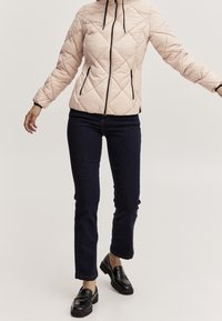 Persona con una giacca trapuntata rosa chiaro, jeans slim blu scuro e mocassini neri spessi, in piedi con le braccia leggermente allargate.