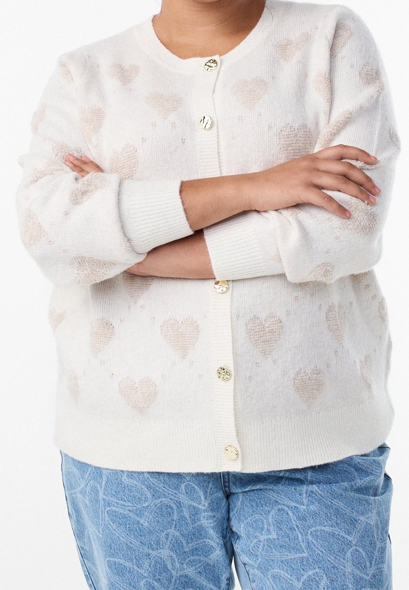 Personne les bras croisés portant un cardigan blanc avec des motifs de cœurs beiges et un jean bleu avec des contours de cœurs blancs.