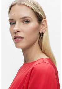 Punainen satiinipaita pyöreällä kaula-aukolla, sileä pinta ja virtaava muotoilu. Hopeiset pisaramuotoiset korvakorut korostavat kokonaisuutta.