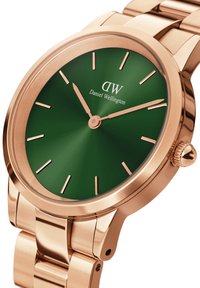 Daniel Wellington zaļš pulkstenis ar pulētu rozā zelta korpusu un rokassprādzi, ar minimālistiskiem stundu atzīmēm un kroni pulksteņa rādītāja pozīcijā, kas norāda 3.