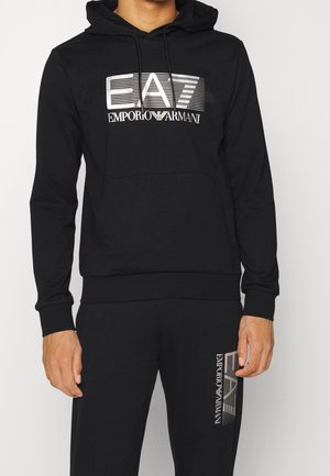 Mężczyzna w czarnej bluzie z kapturem i spodniach z srebrnym logo EA7 Emporio Armani na piersi i lewym udzie.