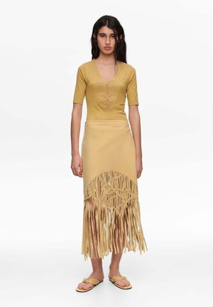 Mujer de pie con un top amarillo mostaza con detalles de ojales y una falda a juego con flecos y recortes geométricos.