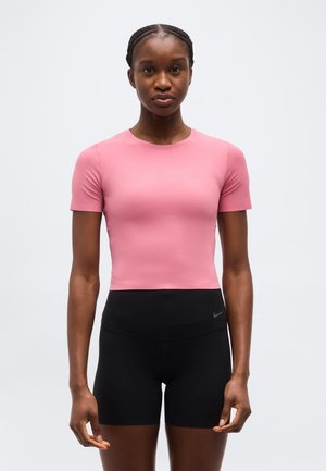 Kobieta z zaplecionymi włosami, ubrana w dopasowany różowy top z krótkim rękawem i czarne sportowe szorty z wysokim stanem na jednolitym tle.