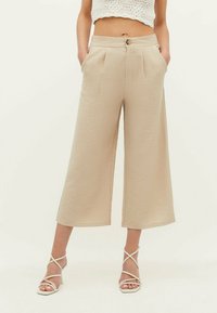 Pantalons larges beiges en tissu texturé, avec des plis à l'avant, fermeture à bouton et poches latérales. Associés à des sandales blanches à lanières.