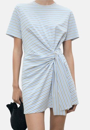 Vrouw draagt een blauw-wit gestreepte jurk met korte mouwen en een knoopdetail aan de voorkant, houdt een kleine zwarte gevlochten handtas in haar linkerhand.