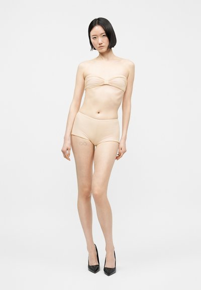 Γυναίκα με κοντά μαύρα μαλλιά που φοράει μπεζ τοπ bandeau και ταιριαστό σορτς με ψηλή μέση, στέκεται φορώντας μαύρα μυτερά ψηλοτάκουνα, σε λευκό φόντο.