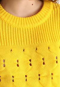 Pull jaune en maille avec un col côtelé et une texture à motif ondulé, agrémenté de petits œillets sur tout le tissu.