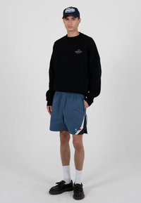 Junger Mann trägt ein schwarzes Sweatshirt, blaue Sportshorts, weiße Kniestrümpfe, schwarze Quastenloafers und eine marineblaue Kappe.