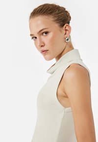 Cream sleeveless top with a high collar and ribbed texture, featuring a fitted design and minimalistic styling. Silver earring accent.