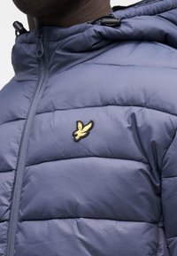 Navy blue padded jacket with a zip closure, featuring an embroidered yellow bird logo on the chest and adjustable drawstring hood.