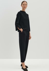 Haut noir à manches longues avec col haut et fentes latérales, associé à un pantalon noir fuselé et des chaussures plates noires, texture de tissu lisse.