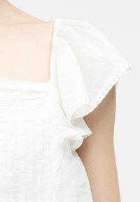 Λευκό μπλουζάκι με τετράγωνη λαιμόκοψη και κοντά μανίκια με δαντελένιο περίγραμμα στο πάνω μέρος του κορμού ενός ατόμου.