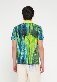 Kortærmet polo-shirt med et levende, abstrakt mønster i blå og neongrøn, med en ensfarvet gul krave og blød stoftekstur.