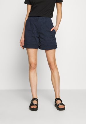 Personne portant un short bleu marine à ourlet roulé, un haut noir à manches courtes et des sandales noires avec des lanières et des œillets argentés, debout avec une main dans la poche.
