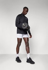 Egy férfi súlyzóval emel, fekete sportdzsekit, fehér rövidnadrágot, fehér zoknit és fekete sportcipőt visel, egy sima háttér előtt.