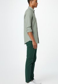 Hombre de perfil vistiendo una camisa de manga larga gris claro, pantalones verde oscuro y zapatillas blancas contra un fondo blanco.