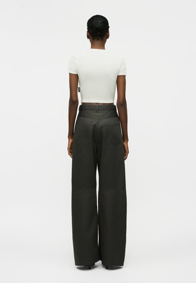 Γυναίκα με κοντά μαλλιά, φορώντας ένα εφαρμοστό λευκό κοντό τοπ και ψηλόμεσα, φαρδιά μαύρα παντελόνια, στέκεται γυρισμένη με την πλάτη σε ένα απλό φόντο.